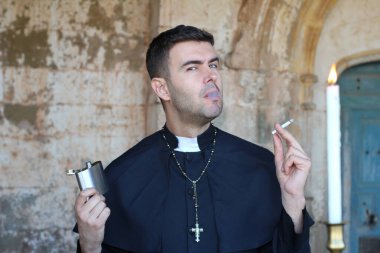 Rahip sigara içiyor ve alkol içiyor. Kilise arka planında yanan mumla poz veriyor.