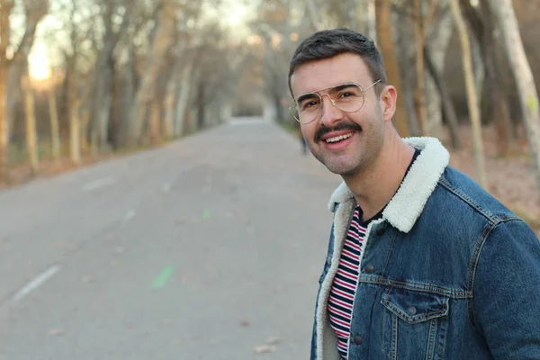 Young man with mustache wearing denim jacket and eyeglasses in autumnal park