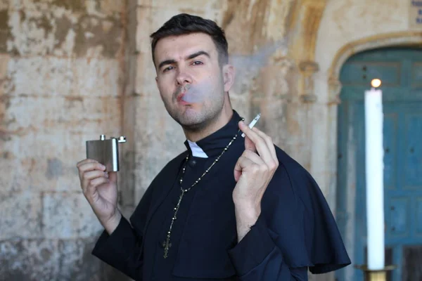 Priest smoking cigarette and drinking alcohol posing on church background