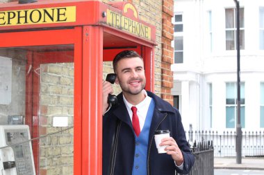 Yakışıklı genç işadamının yakın plan portresi. Elinde kağıt bardak kahve ile sokakta telefon kulübesinde konuşuyor.