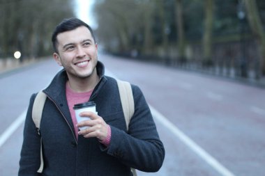 Sırt çantalı ve kağıttan bir fincan kahveyle yolda duran sıradan giysili yakışıklı genç adamın yakın plan portresi.