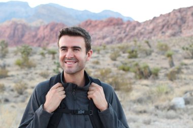 Sırt çantalı gülen adam doğal arka planda poz veriyor.