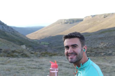 Yeşil çayır üzerinde bir şişe suyla arka planda tepeler olan yakışıklı koşucunun yakın plan portresi.