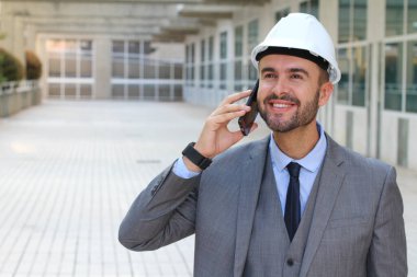 close-up portrait of handsome young businessman in hard hat talking by phone on street clipart