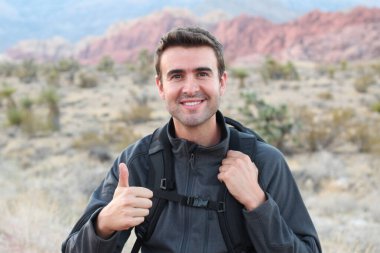 Sırt çantalı gülümseyen adam doğal arka planda baş parmağını gösteriyor.