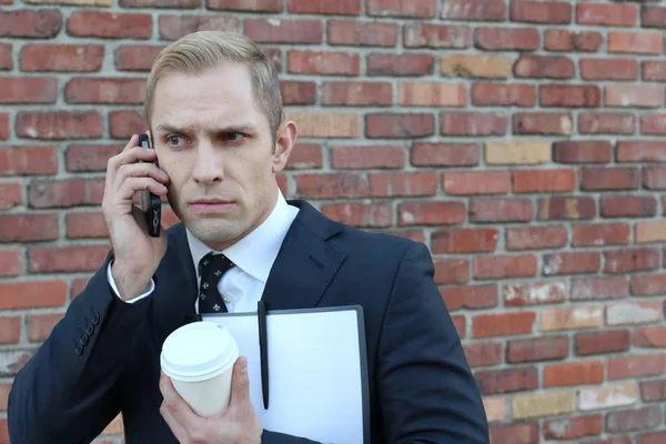 Yakışıklı, yetişkin iş adamının dosyasıyla ve bir fincan kahveyle sokak duvarının önünde telefonla konuşurken yakın plan fotoğrafı.
