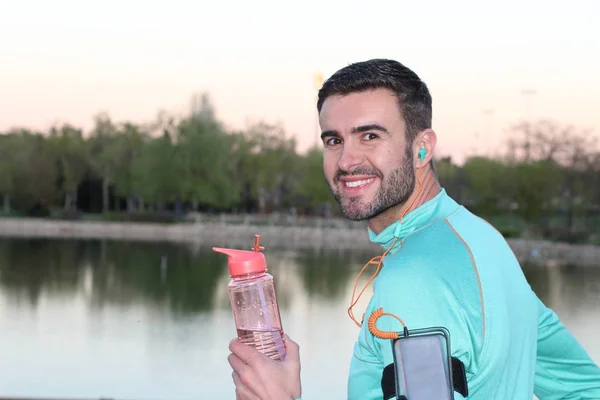 close-up portrait of handsome young jogger with smartphone, sport bottle and earphones in front of lake in park