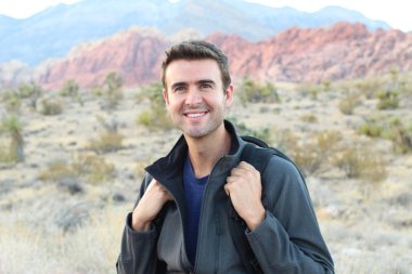 Sırt çantalı gülen adam doğal arka planda poz veriyor.