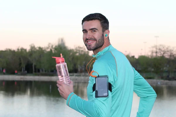 close-up portrait of handsome young jogger with smartphone, sport bottle and earphones in front of lake in park