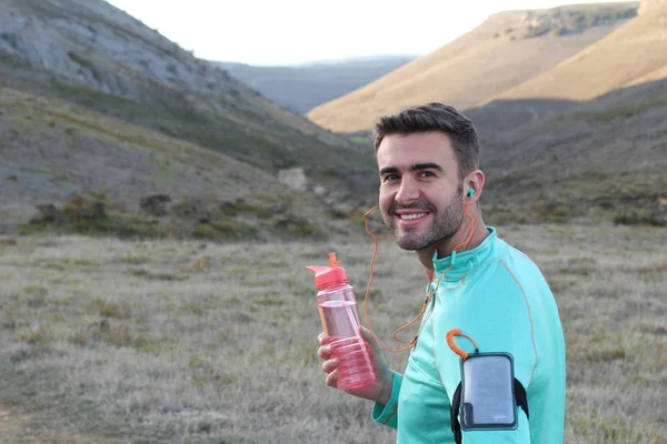 close-up portrait of handsome young jogger with bottle of water on green meadow with hills on background