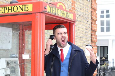 Yakışıklı genç işadamının yakın plan portresi. Elinde kağıt bardak kahve ile sokakta telefon kulübesinde konuşuyor.