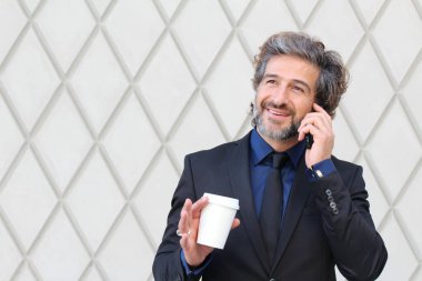Siyah takım elbiseli yakışıklı genç iş adamının yakın plan fotoğrafı. Elinde telefonla konuşan bir fincan kağıt kahve.