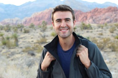 Sırt çantalı gülen adam doğal arka planda poz veriyor.