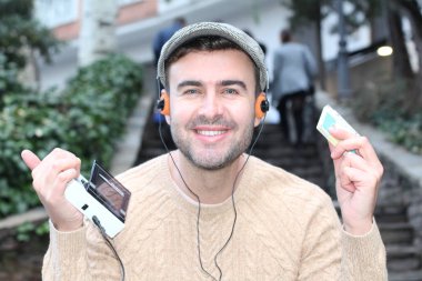 Yakından çekilmiş bir portre, günlük giysiler içindeki yakışıklı genç adam klasik kulaklıklarla müzik dinliyor ve şehir sokaklarında oyuncu.