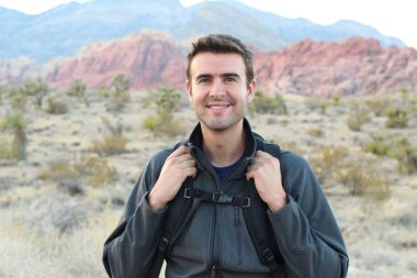 Sırt çantalı gülen adam doğal arka planda poz veriyor.