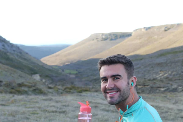 close-up portrait of handsome young jogger with bottle of water on green meadow with hills on background