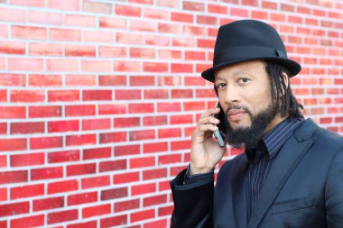 Siyah takım elbiseli, kırmızı tuğla duvarın önünde şapka ve rastalı genç adamın yakın plan portresi.