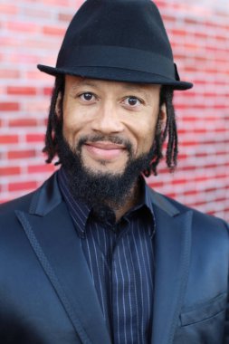 Siyah takım elbiseli, kırmızı tuğla duvarın önünde şapka ve rastalı genç adamın yakın plan portresi.