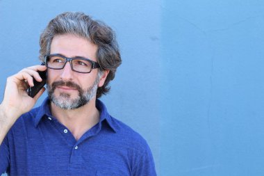 Mavi duvarın önünde telefonla konuşan sıradan giyinmiş yakışıklı yetişkin bir adamın yakın plan portresi.