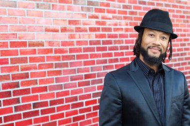 Siyah takım elbiseli, kırmızı tuğla duvarın önünde şapka ve rastalı genç adamın yakın plan portresi.