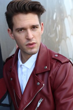 Bad boy concept. Portrait of brutal young man with short hair wearing red leather jacket, jeans and posing over urban background. Hand in pockets. Hipster style. Copy-space. Outdoor shot