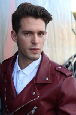 Bad boy concept. Portrait of brutal young man with short hair wearing red leather jacket, jeans and posing over urban background. Hand in pockets. Hipster style. Copy-space. Outdoor shot