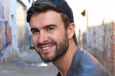 Genç, çekici bir hippi ve modaya uygun görünüşlü bir adam gülümsüyor. Mutlu pozlar veriyor. Gayri resmi giyiniyor, beysbol şapkası ve sakal takıyor.