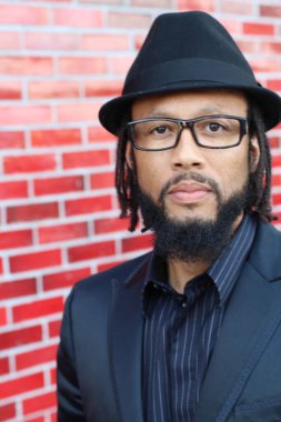 Siyah takım elbiseli, kırmızı tuğla duvarın önünde şapka ve rastalı genç adamın yakın plan portresi.