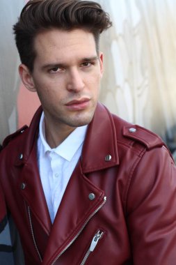 Bad boy concept. Portrait of brutal young man with short hair wearing red leather jacket, jeans and posing over urban background. Hand in pockets. Hipster style. Copy-space. Outdoor shot
