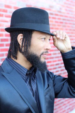 Siyah takım elbiseli, kırmızı tuğla duvarın önünde şapka ve rastalı genç adamın yakın plan portresi.