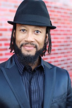 Siyah takım elbiseli, kırmızı tuğla duvarın önünde şapka ve rastalı genç adamın yakın plan portresi.