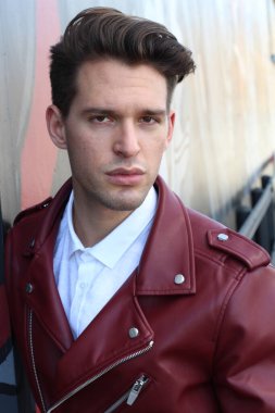 Bad boy concept. Portrait of brutal young man with short hair wearing red leather jacket, jeans and posing over urban background. Hand in pockets. Hipster style. Copy-space. Outdoor shot