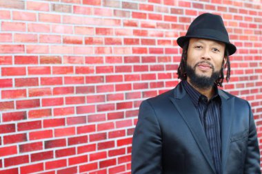 Siyah takım elbiseli, kırmızı tuğla duvarın önünde şapka ve rastalı genç adamın yakın plan portresi.