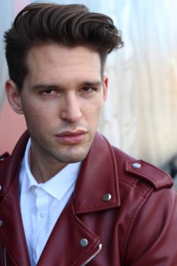 Bad boy concept. Portrait of brutal young man with short hair wearing red leather jacket, jeans and posing over urban background. Hand in pockets. Hipster style. Copy-space. Outdoor shot