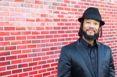 Siyah takım elbiseli, kırmızı tuğla duvarın önünde şapka ve rastalı genç adamın yakın plan portresi.