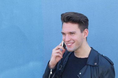 Handsome serious beauty male model portrait wearing leather jacket talking on phone, young guy over blue background