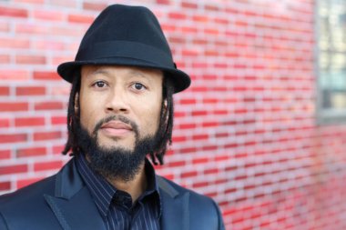 Siyah takım elbiseli, kırmızı tuğla duvarın önünde şapka ve rastalı genç adamın yakın plan portresi.