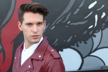 Bad boy concept. Portrait of brutal young man with short hair wearing red leather jacket, jeans and posing over urban background. Hand in pockets. Hipster style. Copy-space. Outdoor shot