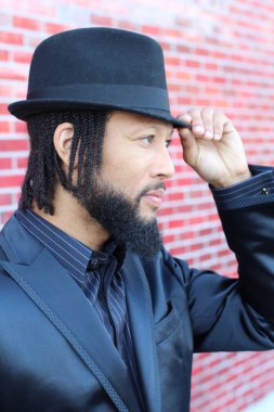 Siyah takım elbiseli, kırmızı tuğla duvarın önünde şapka ve rastalı genç adamın yakın plan portresi.
