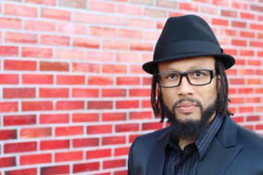 Siyah takım elbiseli, kırmızı tuğla duvarın önünde şapka ve rastalı genç adamın yakın plan portresi.