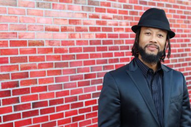 Siyah takım elbiseli, kırmızı tuğla duvarın önünde şapka ve rastalı genç adamın yakın plan portresi.