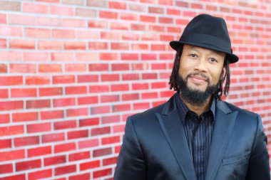 Siyah takım elbiseli, kırmızı tuğla duvarın önünde şapka ve rastalı genç adamın yakın plan portresi.