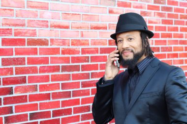 Siyah takım elbiseli, kırmızı tuğla duvarın önünde şapka ve rastalı genç adamın yakın plan portresi.