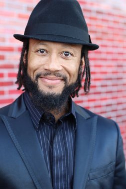 Siyah takım elbiseli, kırmızı tuğla duvarın önünde şapka ve rastalı genç adamın yakın plan portresi.