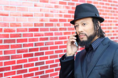 Siyah takım elbiseli, kırmızı tuğla duvarın önünde şapka ve rastalı genç adamın yakın plan portresi.