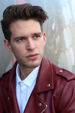 Bad boy concept. Portrait of brutal young man with short hair wearing red leather jacket, jeans and posing over urban background. Hand in pockets. Hipster style. Copy-space. Outdoor shot