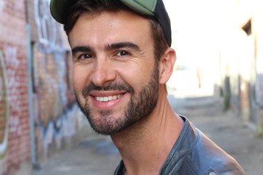 Genç, çekici bir hippi ve modaya uygun görünüşlü bir adam gülümsüyor. Mutlu pozlar veriyor. Gayri resmi giyiniyor, beysbol şapkası ve sakal takıyor.