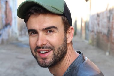 Genç, çekici bir hippi ve modaya uygun görünüşlü bir adam gülümsüyor. Mutlu pozlar veriyor. Gayri resmi giyiniyor, beysbol şapkası ve sakal takıyor.