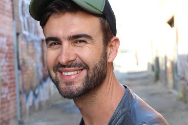 Genç, çekici bir hippi ve modaya uygun görünüşlü bir adam gülümsüyor. Mutlu pozlar veriyor. Gayri resmi giyiniyor, beysbol şapkası ve sakal takıyor.