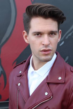 Bad boy concept. Portrait of brutal young man with short hair wearing red leather jacket, jeans and posing over urban background. Hand in pockets. Hipster style. Copy-space. Outdoor shot